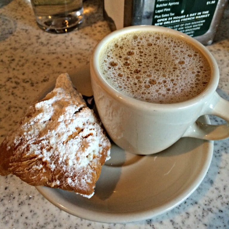 a cup of steaming cafe au lait and a beignet on a saucer